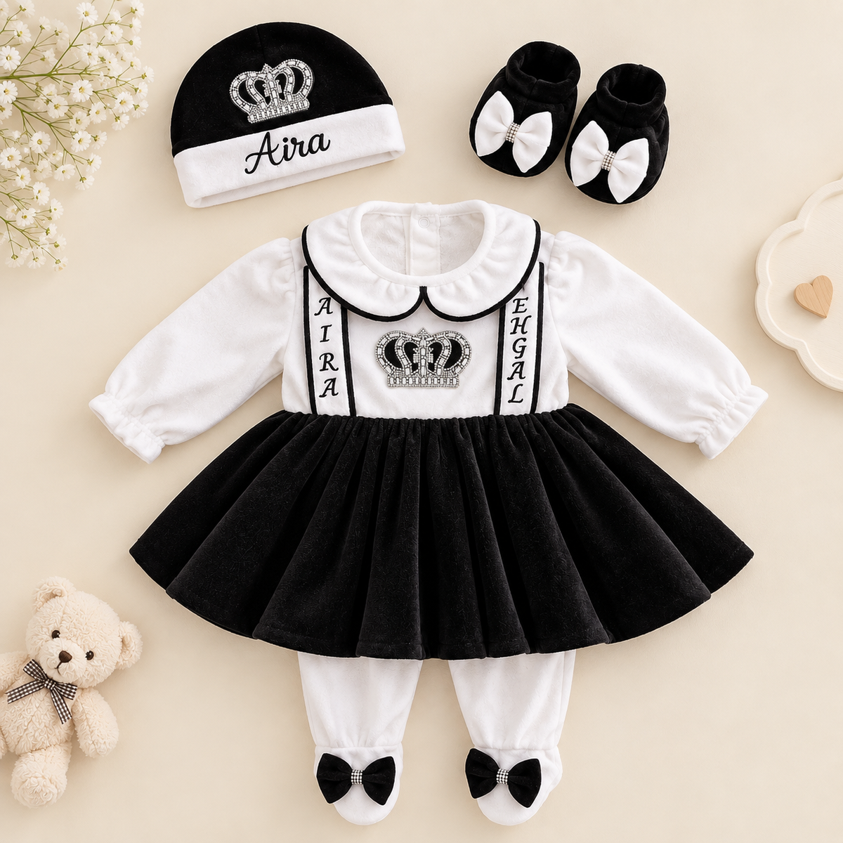A baby girl's two-piece outfit set featuring a black and white long-sleeve dress with a black tutu skirt, white Peter Pan collar, and 'AIRA SEHGAL' text with a crown graphic. The set includes a matching beanie with 'Aira' and a crown, black booties with white bows, white leggings with black bow feet, a teddy bear, and baby flowers on a beige background.