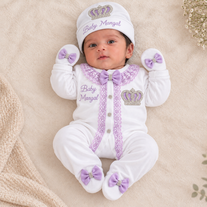 A baby lying on a soft beige blanket, wearing a white footed onesie with lavender lace trim, lavender bow details, and a matching white beanie. The outfit has 'Baby Mangal' embroidered on the chest and a lavender crown graphic on the shoulder.