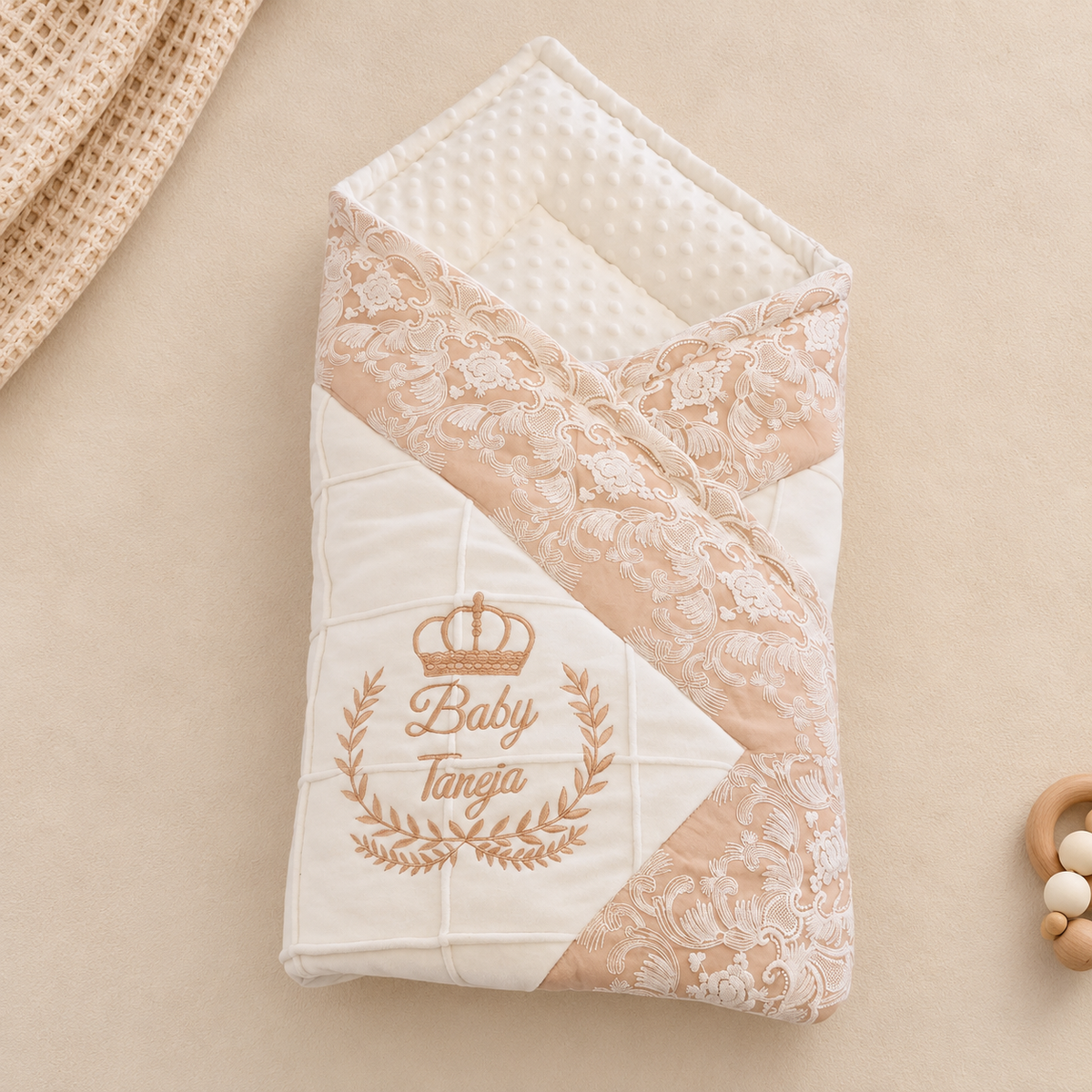 A folded baby swaddle blanket with a cream and beige design, featuring a lace pattern, a crown graphic, and the personalized text 'Baby Tangja' in gold embroidery, displayed on a neutral beige background.