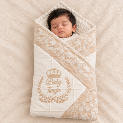 A sleeping newborn baby wrapped in a cream and beige quilted swaddle blanket with floral lace trim and embroidered 'Baby Tangia' text with a crown, laid on a textured beige blanket.