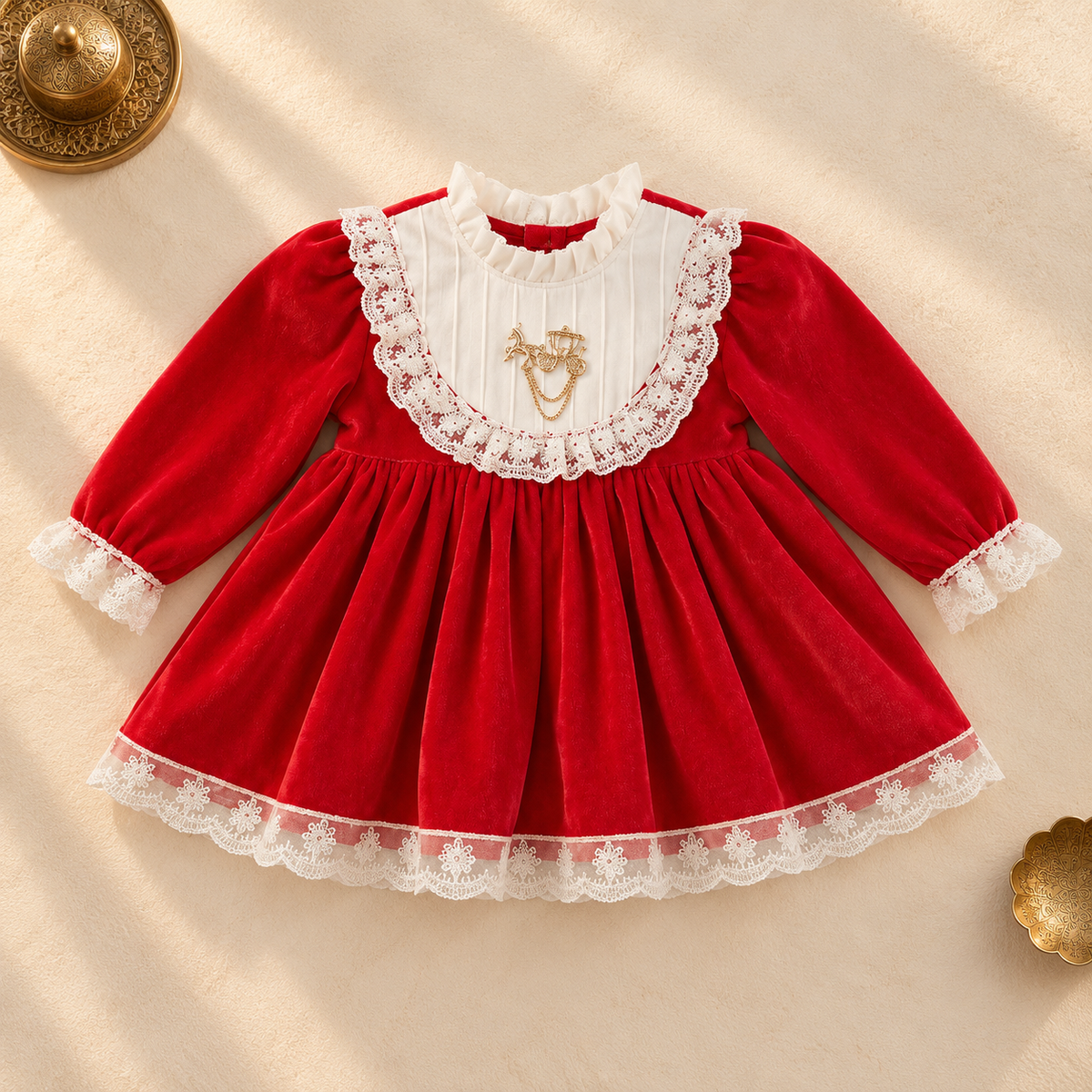 A baby girl's long-sleeve dress in red velvet with a white ruffled collar, white lace trim, and a gold embroidered monogram on the chest, laid flat on a beige background with decorative gold props.