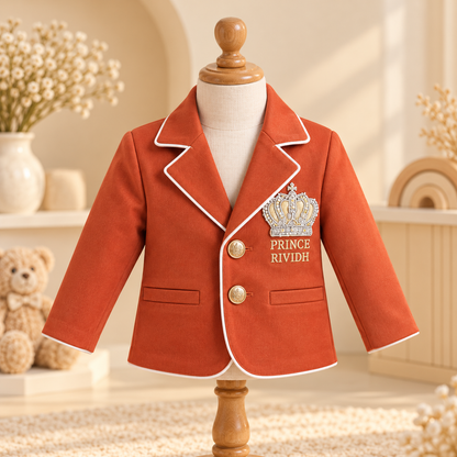A toddler boy's orange-red blazer with white piping and a gold crown patch reading 'PRINCE RIVIDH' on the chest, displayed on a wooden mannequin torso in a warm, styled nursery setting.