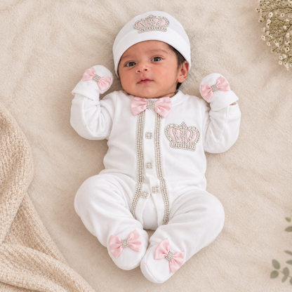 A baby wearing a white footed onesie with pink bow details, rhinestone crown embellishments, and a matching white bonnet, lying on a soft beige blanket.