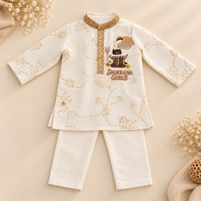 A baby's traditional Indian outfit consisting of a cream-colored long-sleeve kurta and matching pants, decorated with gold floral patterns and a brown graphic of Shri Krishna on the chest. The outfit is displayed flat on a beige background with decorative dried flowers.