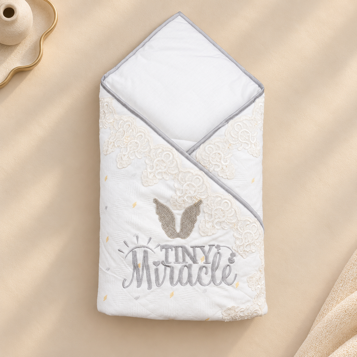 A folded white baby blanket with a gray hood and trim, decorated with beige lace-like embroidery, a gray angel wing motif, and the words 'TINY Miracle' in gray and beige, shown on a warm beige fabric background.