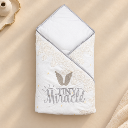 A folded white baby blanket with a gray hood and trim, decorated with beige lace-like embroidery, a gray angel wing motif, and the words 'TINY Miracle' in gray and beige, shown on a warm beige fabric background.