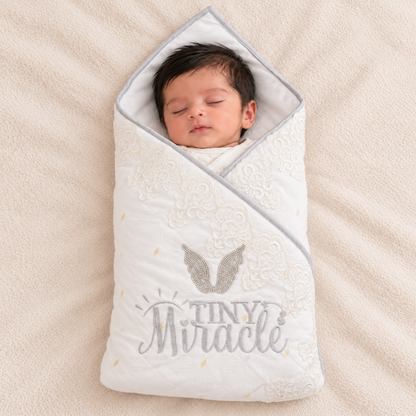 A sleeping newborn baby swaddled in a white quilted wrap blanket with gray embroidered text reading 'TINY Miracle' and a gray angel wing design, lying on a beige textured blanket.