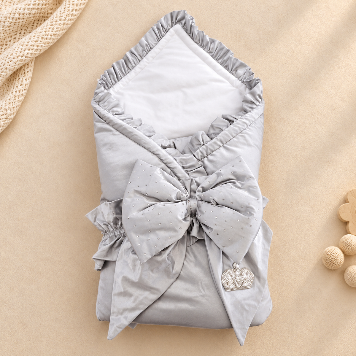 A folded baby swaddle blanket in light gray with a large bow, ruffled edges, and a small silver heart charm, photographed on a warm beige background with soft decorative props.