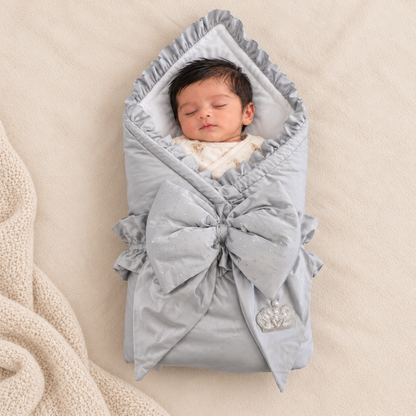 A sleeping newborn baby wrapped in a light gray swaddle blanket with a large bow and ruffled trim, lying on a beige textured blanket.