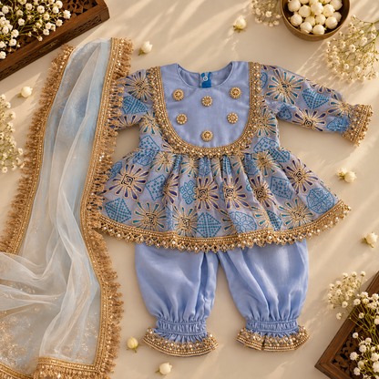 A light blue traditional outfit set for a baby, featuring an embroidered peplum top, matching pants, and a sheer dupatta, arranged in a styled flat-lay with decorative flowers and wooden trays.
