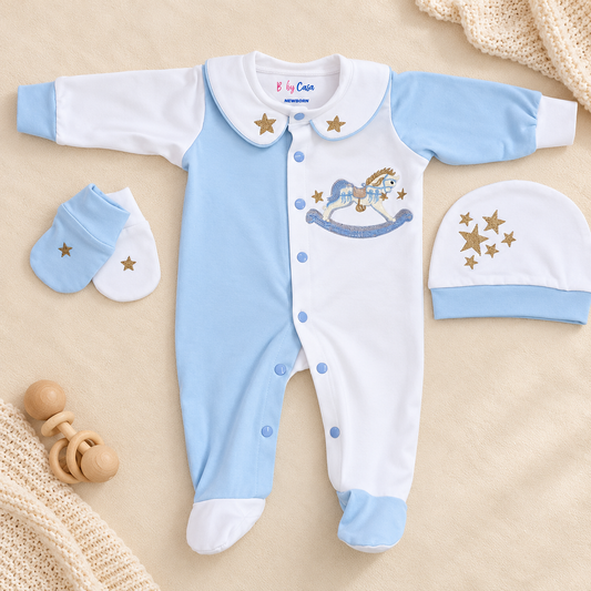 A baby footed sleeper set in light blue and white, featuring a central rocking horse graphic with gold stars. The set includes a matching sleeper hat and a pair of booties, all laid out on a beige fabric background with soft knit blankets and a wooden teether.