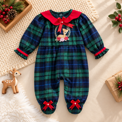 A baby holiday-themed plaid jumpsuit laid flat on a cream knit blanket with Christmas props. The jumpsuit features a green and navy blue tartan pattern, a bright red collar with a red bow, red trim on the cuffs and ankles, and a central embroidered deer design.