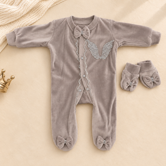 A baby sleep suit in soft taupe-gray velvet with silver rhinestone wing and bow details, shown flat-laid with matching booties on a beige fabric background.