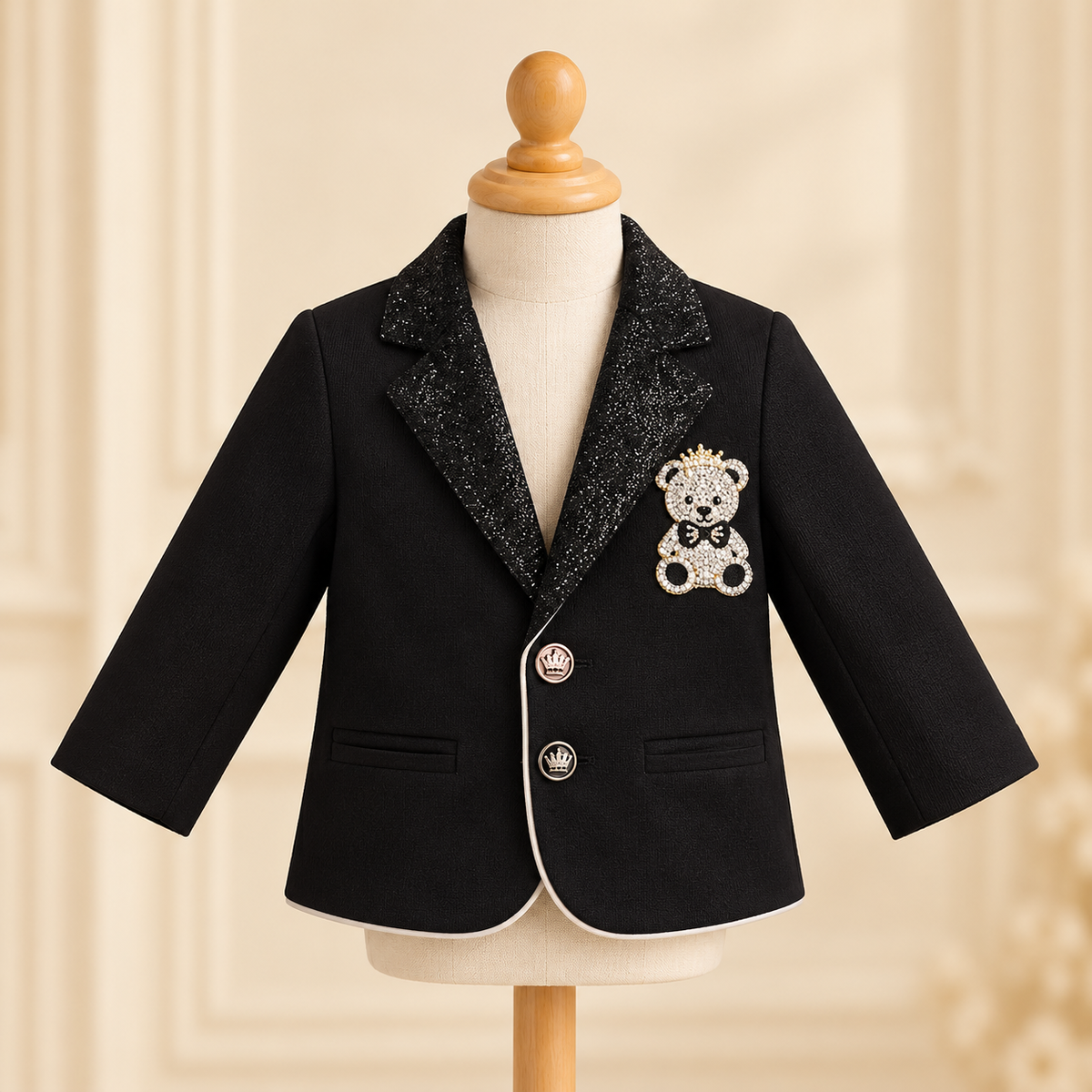 A black baby boy's blazer with a sparkling black glitter lapel, featuring a teddy bear brooch on the chest and white piping along the front edge, displayed on a mannequin torso.