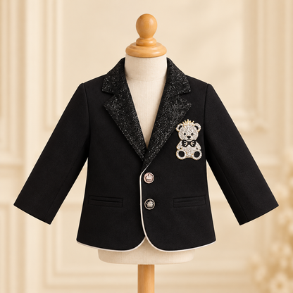 A black baby boy's blazer with a sparkling black glitter lapel, featuring a teddy bear brooch on the chest and white piping along the front edge, displayed on a mannequin torso.