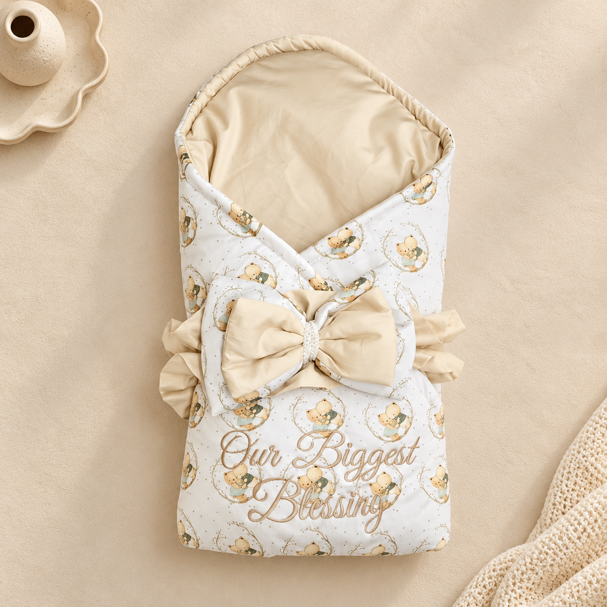 A baby wrap blanket or swaddle with a cream and beige teddy bear pattern, featuring a large matching bow, ruffled trim, and the words 'Our Biggest Blessing' printed on the front. The blanket is styled on a beige textured background with a small ceramic dish in the corner.