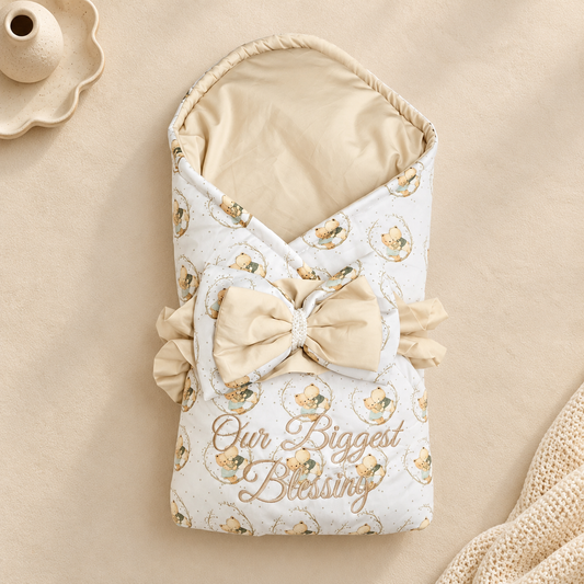 A baby wrap blanket or swaddle with a cream and beige teddy bear pattern, featuring a large matching bow, ruffled trim, and the words 'Our Biggest Blessing' printed on the front. The blanket is styled on a beige textured background with a small ceramic dish in the corner.