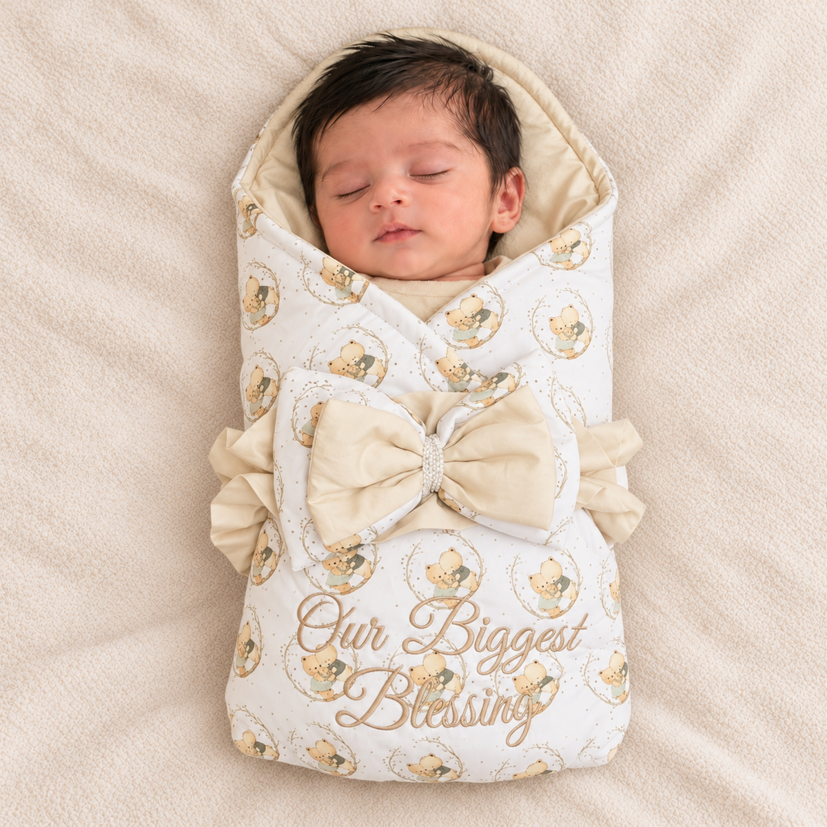 A sleeping newborn baby wrapped in a cream and beige patterned swaddle blanket with a large bow and the words 'Our Biggest Blessing' printed on the front, lying on a soft beige blanket.