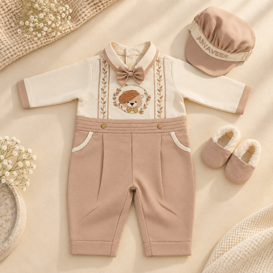 A baby outfit set styled flat-lay on a beige fabric background, including a long-sleeve romper with cream and dusty pink sections, a bow tie, embroidered chest detail, matching cap, and booties. The cap shows the brand name 'ANVAVEER'.