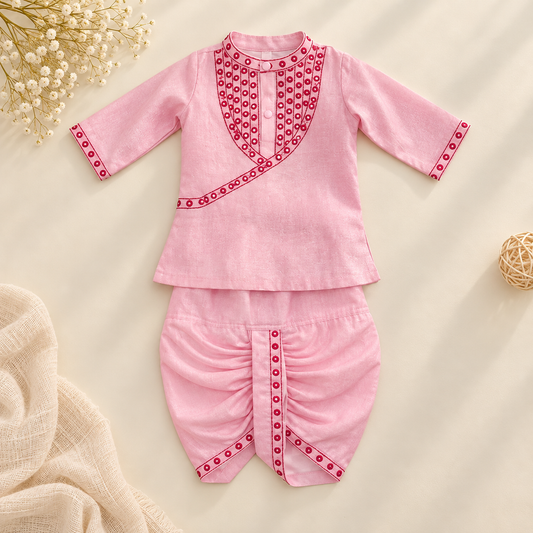A baby outfit set consisting of a pink long-sleeve top and matching pants, decorated with red and white geometric patterns along the neckline, sleeve cuffs, and front center. The set is displayed flat on a light beige background with decorative props including dried flowers, a textured beige cloth, and a small woven ball.