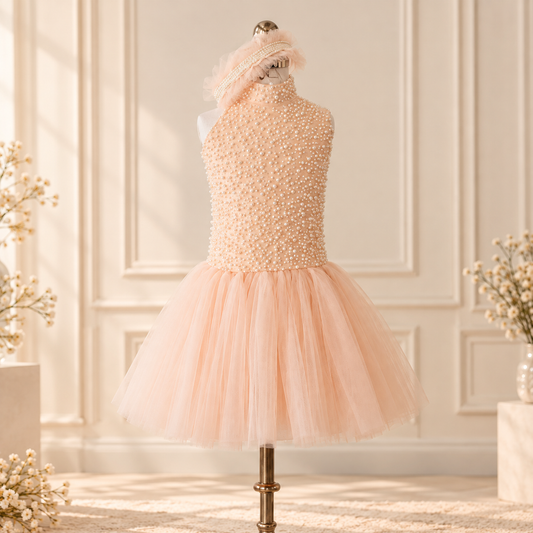 A peach-colored formal dress displayed on a mannequin in an elegant cream-colored room. The dress features a beaded halter-style bodice with pearl-like embellishments and a voluminous tulle skirt, accompanied by a matching feather-trimmed headband.