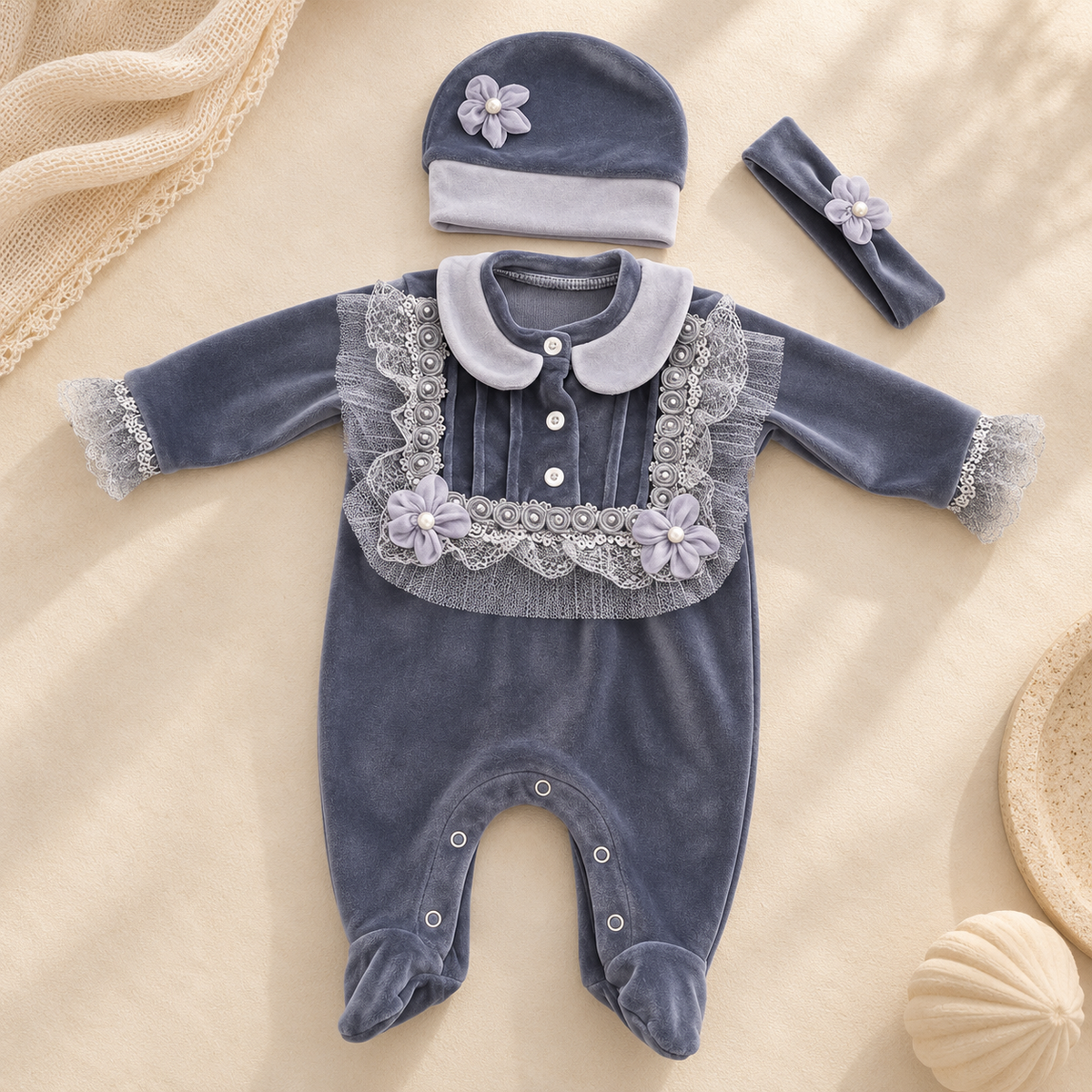 A baby footed one-piece outfit set in dusty blue velvet, featuring a Peter Pan collar, lace trim, and floral appliques. The set includes a matching bonnet and a headband, arranged on a beige fabric background with decorative props.