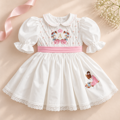 A white baby girl's dress with puff sleeves, a pink waistband, and embroidered details. The bodice features an embroidered monogram 'A' surrounded by flowers and bows, and the skirt has a small embroidered character.