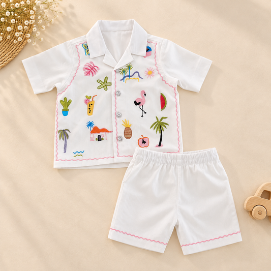 A baby boy's two-piece pajama set featuring a short-sleeve button-up shirt and matching shorts. The set is white with colorful tropical-themed embroidery (including palm trees, flamingos, pineapples, and cacti) and pink zigzag trim.