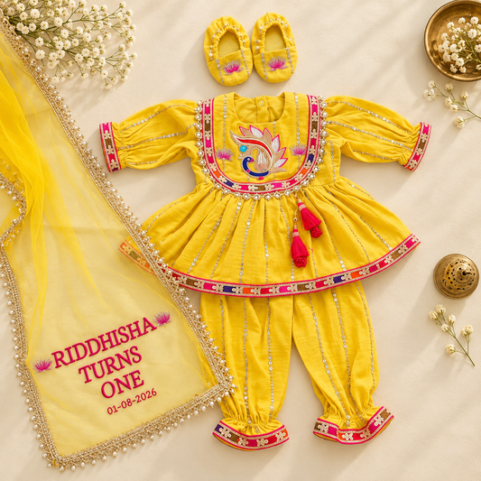 A yellow baby girl's traditional Indian outfit set laid flat on a cream fabric background, including a kurta, pants, matching yellow shoes, and a dupatta. The kurta has colorful embroidery on the chest, silver mirror work along the borders, and pink tassel details.
