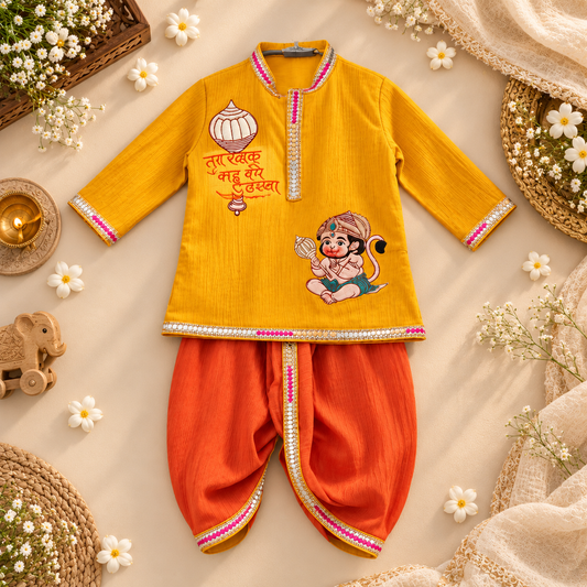 A baby outfit set consisting of a mustard yellow long-sleeve kurta with red and gold embroidery and a cartoon monkey graphic, paired with bright orange pants featuring pink and gold trim, laid flat on a beige fabric background decorated with small white flowers and woven mats.