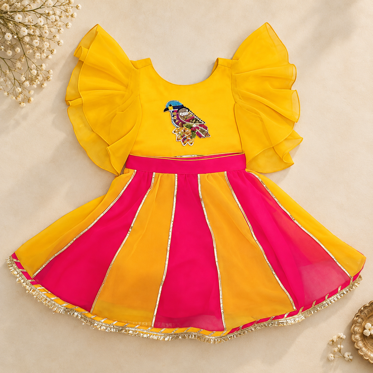 A baby girl's festive dress with a bright yellow bodice featuring ruffled sleeves and a colorful bird embroidery, paired with a pleated skirt in yellow and hot pink with gold trim.