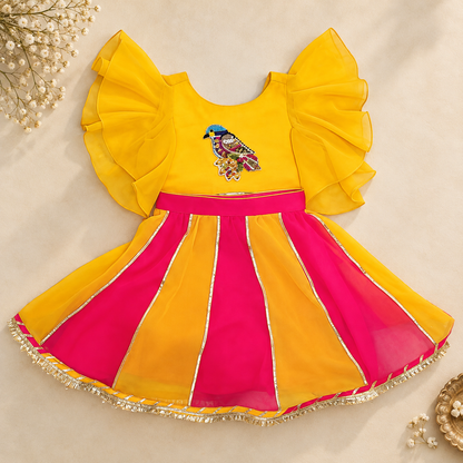 A baby girl's festive dress with a bright yellow bodice featuring ruffled sleeves and a colorful bird embroidery, paired with a pleated skirt in yellow and hot pink with gold trim.